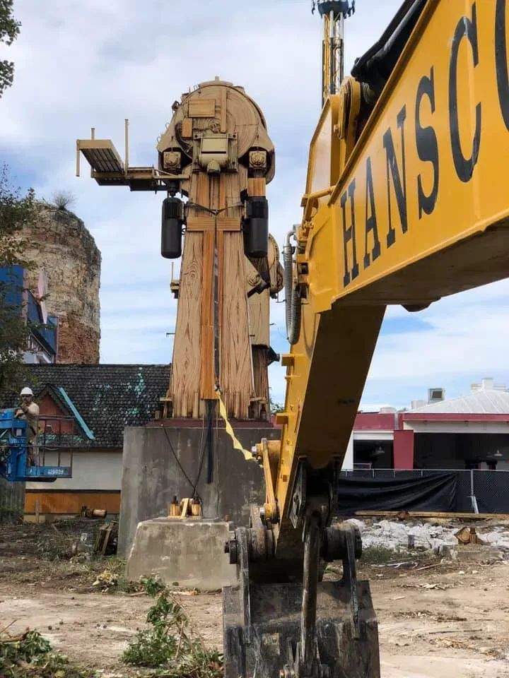 Yellow excavator arm facing a large, tan amusement ride structure. Construction site setting.
