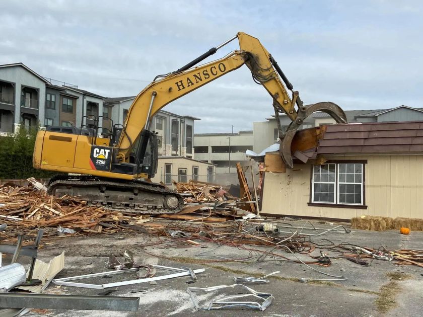 Yellow excavator demolishing a tan building, debris scattered. Buildings in background, overcast sky.