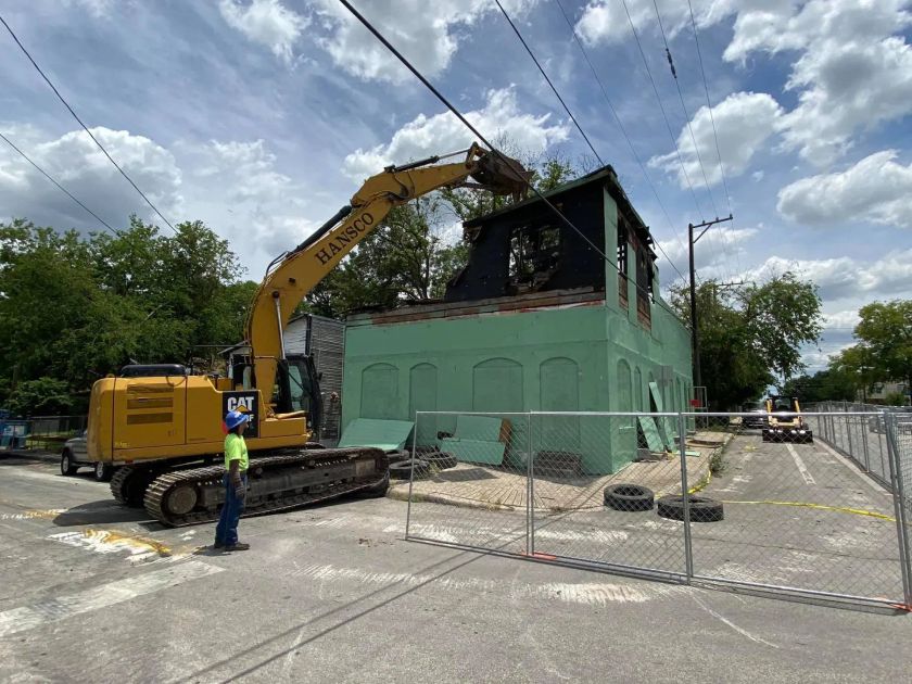 A yellow excavator demolishes a green building, smoke damage visible, under a cloudy sky.