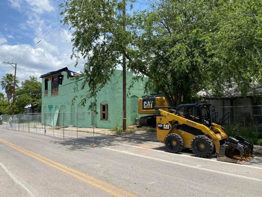A yellow excavator near a light green building with burned roof. Building is behind a chain link fence along a road.
