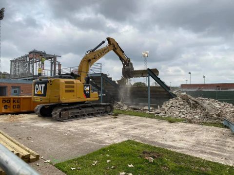Yellow excavator demolishing a building, with debris and a conveyor belt. Cloudy sky.