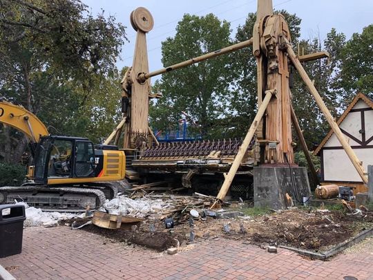 Demolition of a tall amusement park ride. Yellow excavator beside the decaying structure.