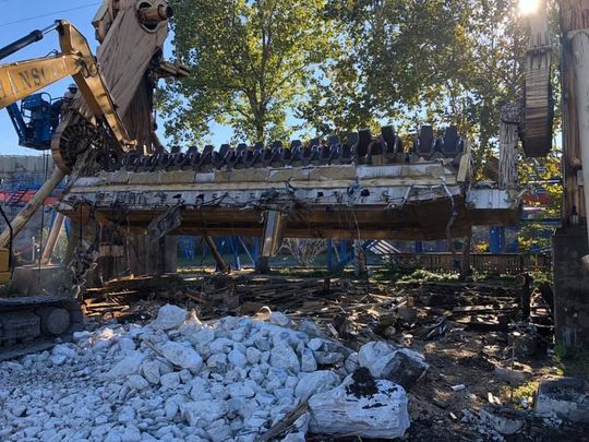 Excavator demolishing a concrete structure, with debris and trees in the background under daylight.