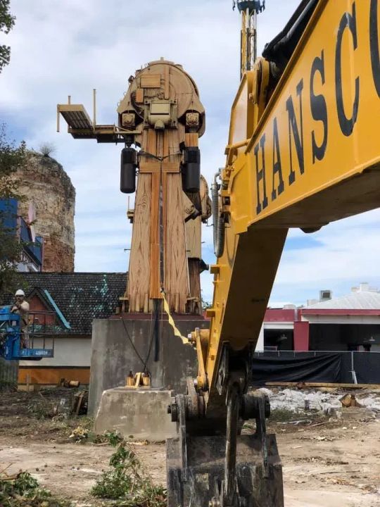 Yellow excavator arm facing a large, tan amusement ride structure. Construction site setting.