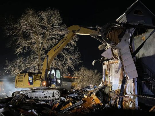 Yellow excavator demolishing a building at night. Debris lit by work lights.