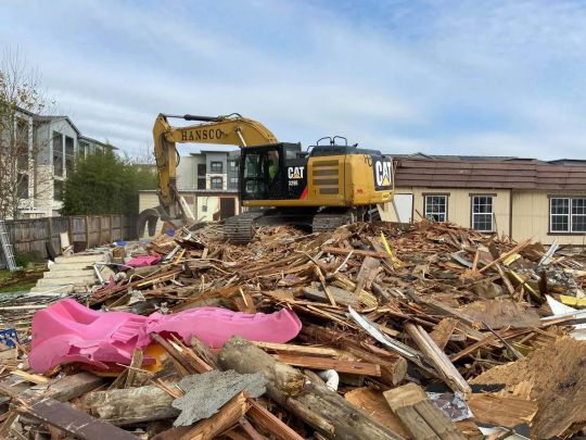 Yellow excavator on a pile of debris, demolishing a building. Cloudy sky in the background.