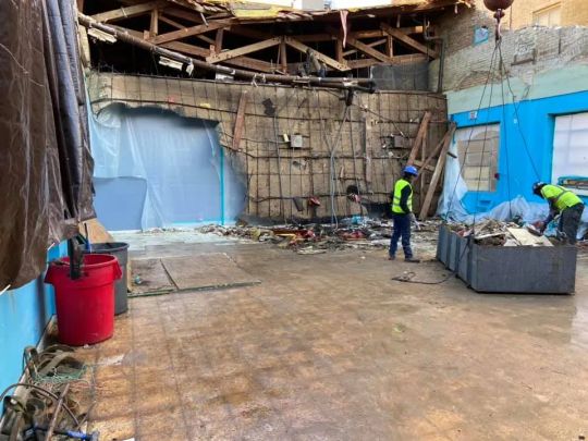 Construction workers inside a partially demolished building. Debris litters the floor; blue walls; exposed ceiling beams.