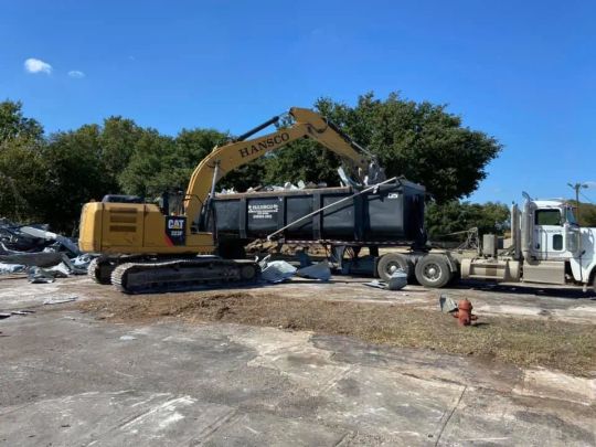 Yellow excavator loading debris into a black dumpster truck on pavement, sunny day.