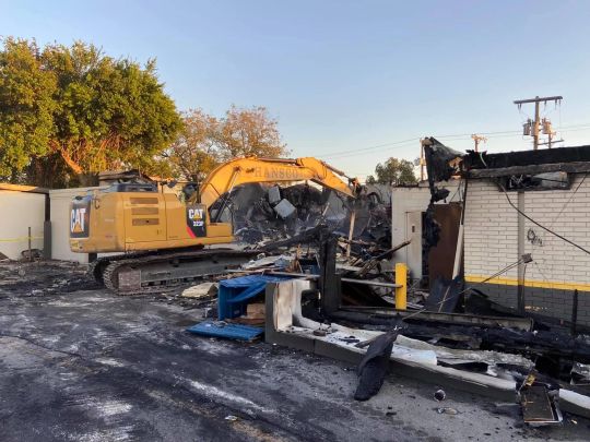 Yellow excavator demolishing a charred building. Outdoor setting, blue sky.