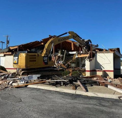 Yellow excavator demolishing a single-story building under a clear sky. Debris surrounds the structure.