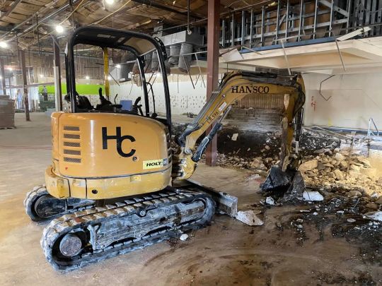 Yellow mini excavator demolishing interior construction. HC logo on side. Construction debris on floor.