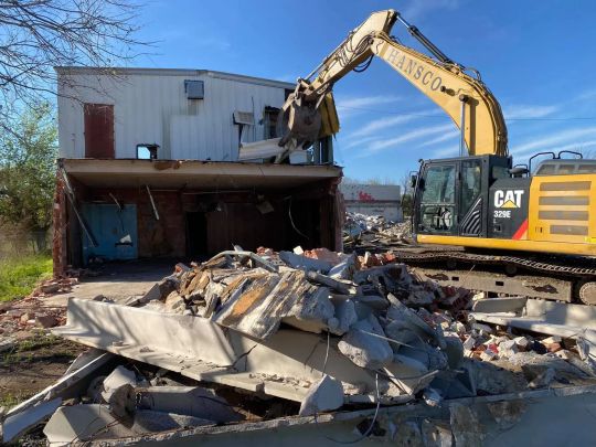 A yellow CAT excavator demolishing a brick building; debris on the ground.