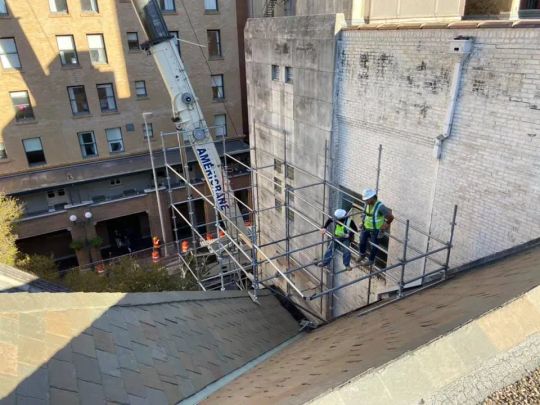Two workers on scaffolding attached to a building's side, crane in the background.