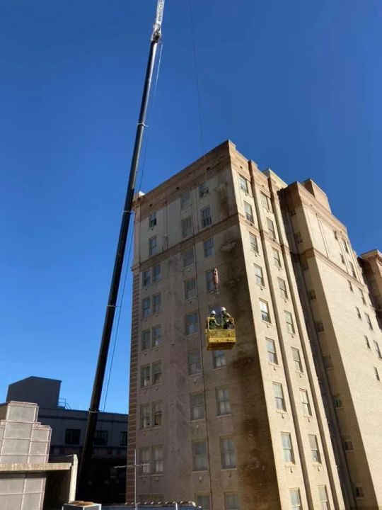 A tall crane lifts a yellow construction basket near a multi-story building. Two workers in the basket. Clear blue sky.