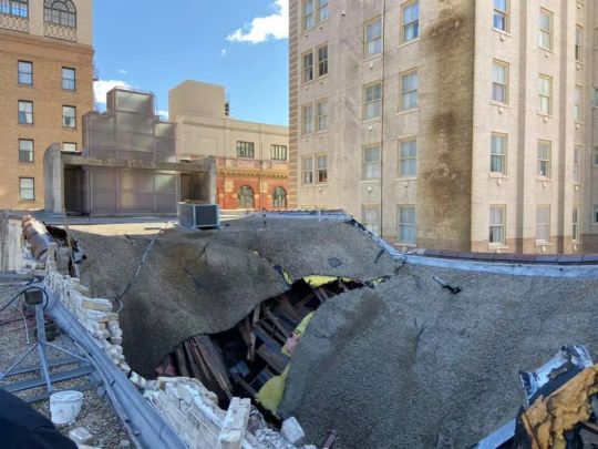 Rooftop partially collapsed, exposing interior. Buildings surround the damaged area on a sunny day.