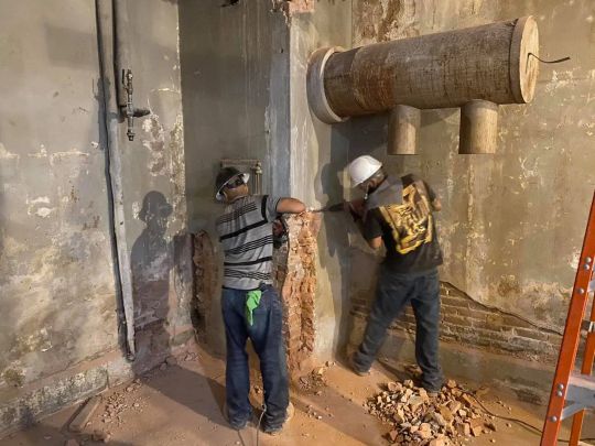 Two workers drilling into a concrete wall with exposed piping. Construction setting.
