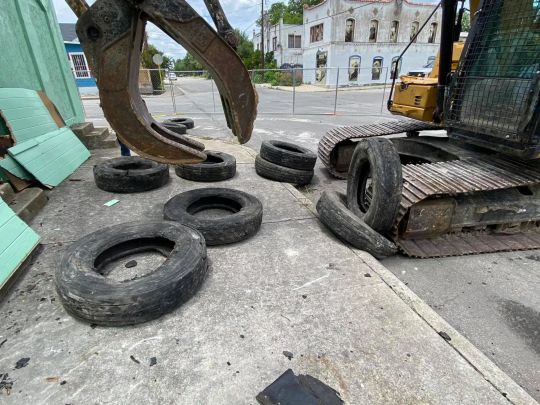 Excavator with claw next to tires on concrete, urban setting, possible demolition in background.