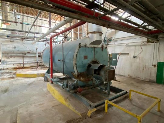 Industrial boiler in an old building, with red and white pipes overhead and a yellow safety barrier.