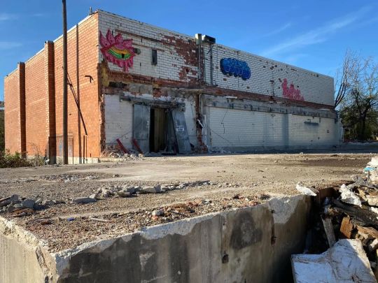 Partially demolished brick building with murals, debris in foreground, blue sky.