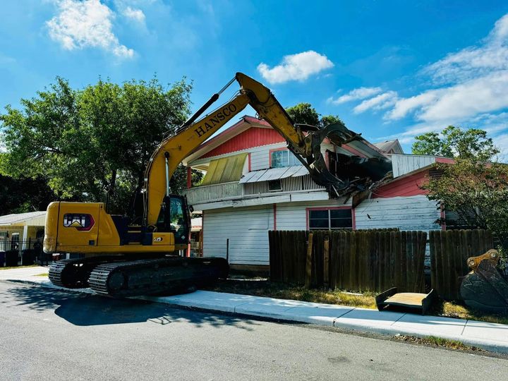 Hansco excavator demolishing a two-story house behind a wooden fence on a residential street